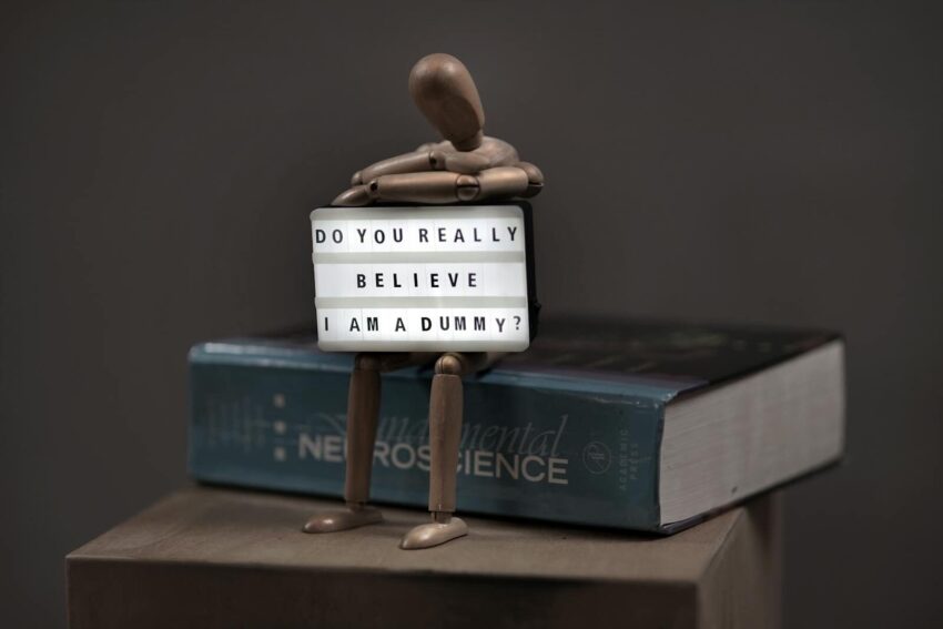 A wooden mannequin poses with a letter board reading 'Do you really believe I am a dummy?' atop a neuroscience book.
